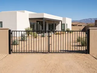 A modern black steel driveway gate of vertical flat bars with narrow gaps in front of a contemporary stucco home